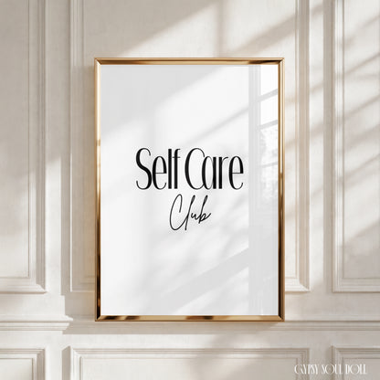 A framed wall art print with the words 'Self Care Club' in black lettering on a white background, set in a gold frame and mounted on a white panelled wall with soft window shadows by Gypsy Soul Doll.