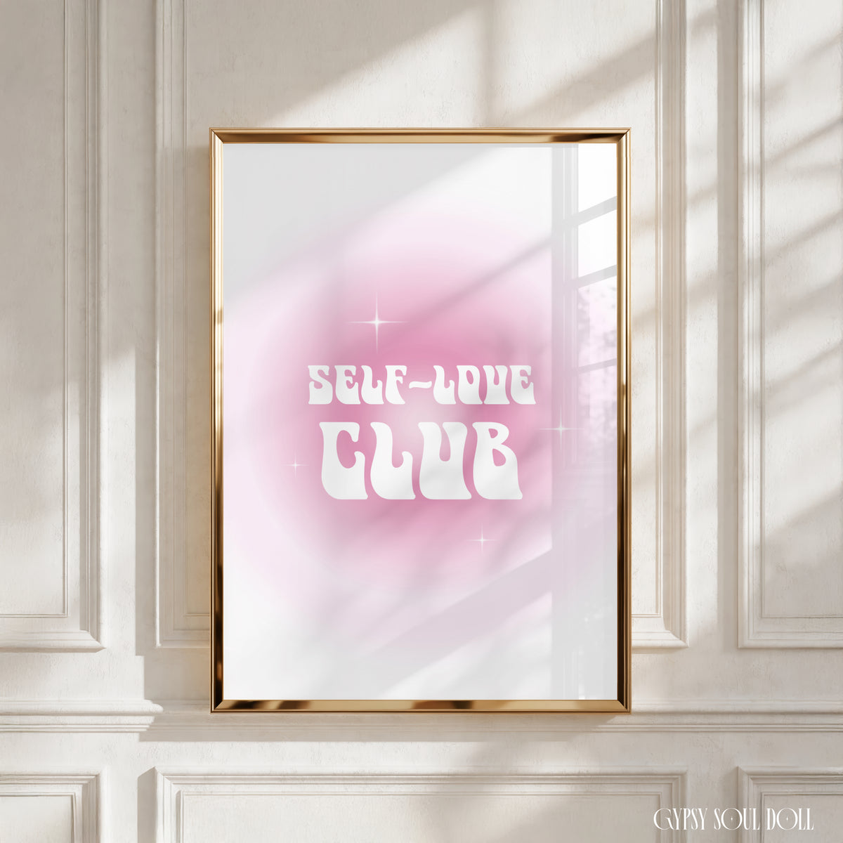 A framed art print with the text 'SELF-LOVE CLUB' in white retro lettering over a pink and white gradient background, displayed in a gold frame on a white panelled wall with soft window shadow light by Gypsy Soul Doll.