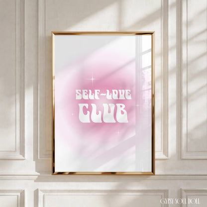 A framed art print with the text 'SELF-LOVE CLUB' in white retro lettering over a pink and white gradient background, displayed in a gold frame on a white panelled wall with soft window shadow light by Gypsy Soul Doll.