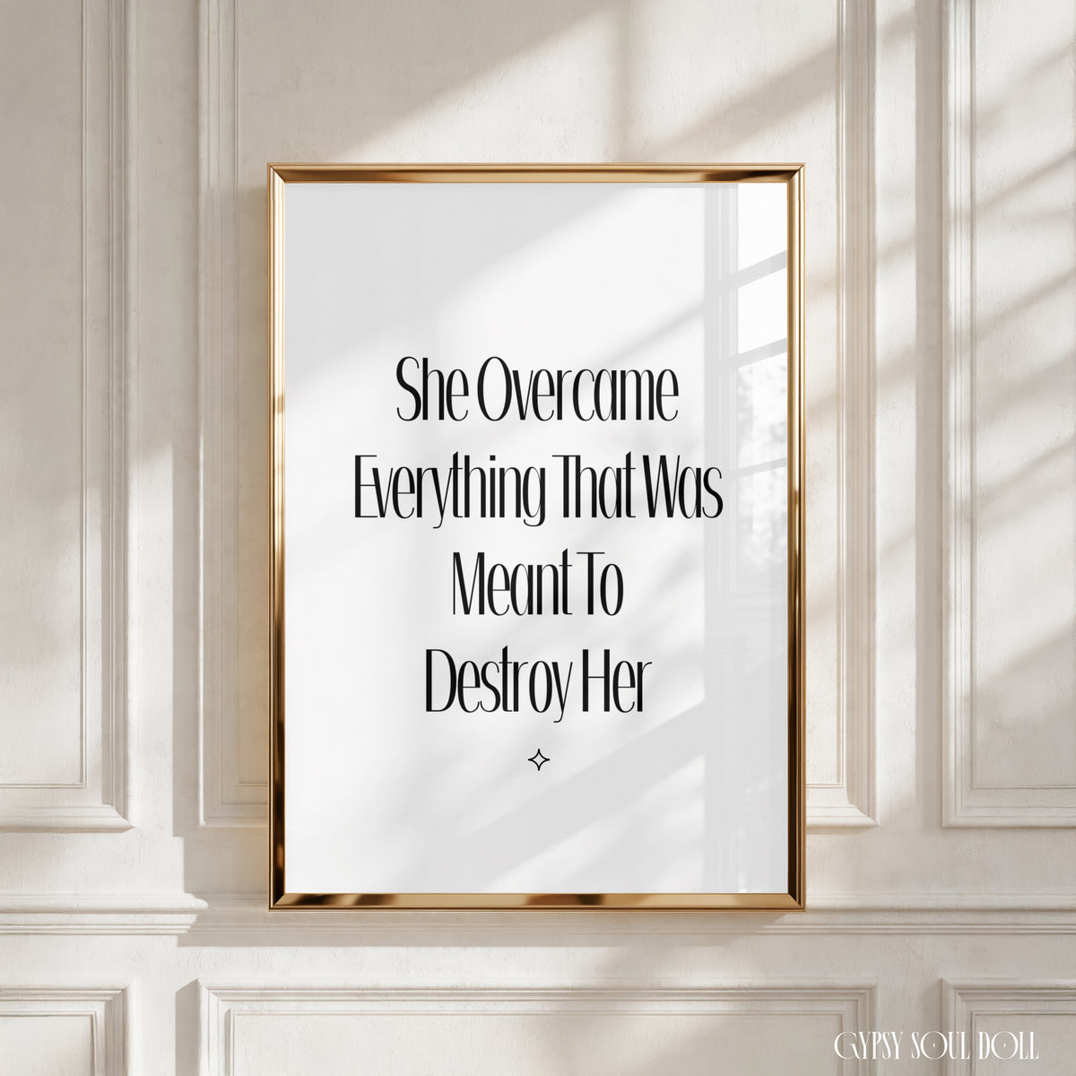 A framed typographic wall art print with black serif text on a white background, set in a thin gold frame and displayed against a white panelled wall with soft window shadows. The print reads ‘ She Overcame Everything That Was Meant to Destroy Her’.