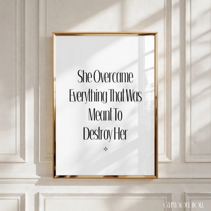 A framed typographic wall art print with black serif text on a white background, set in a thin gold frame and displayed against a white panelled wall with soft window shadows. The print reads ‘ She Overcame Everything That Was Meant to Destroy Her’.