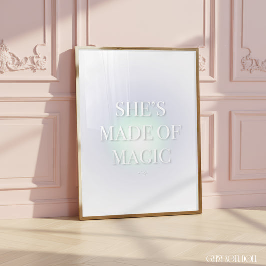 Minimalist A4 celestial print featuring the text SHE'S MADE OF MAGIC in bold white typography over a soft pastel green aura gradient background, leaning in a gold frame on a wooden floor against a pink panelled wall.