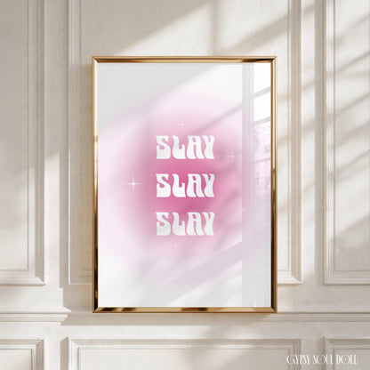 A framed art print featuring the text 'SLAY SLAY SLAY' in white retro-style lettering over a pink and white gradient background, displayed in a gold frame on a white panelled wall with soft window shadows by Gypsy Soul Doll.