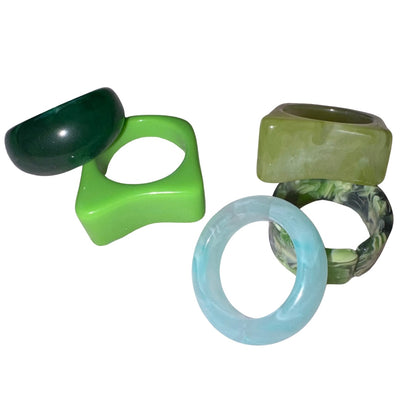 A flat lay studio image displaying a collection of five chunky Y2K style acrylic rings in shades of bright neon lime, forest green, marbled olive, and a translucent icy blue band on a clean white backdrop.