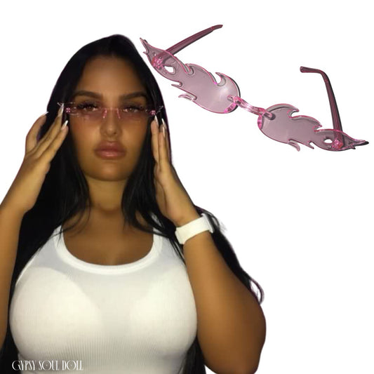 Side-by-side product view featuring a model with long dark hair wearing translucent pink rimless flame-shaped fashion glasses, next to a floating graphic cutout of the pink fire-style shades against a clean white background.