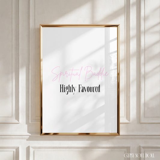 A framed art print with a gold frame hangs on a white panelled wall. The print features the text 'Spiritual Baddie' in pink script and 'Highly Favoured' in black serif font on a white background by Gypsy Soul Doll.