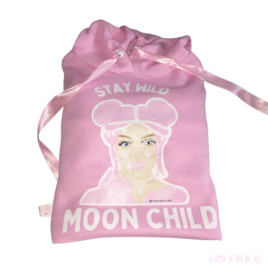 A pink hoodie featuring a graphic of a girl with pink hair and the text 'STAY WILD MOON CHILD' in white, with pink satin ribbon pull strings.