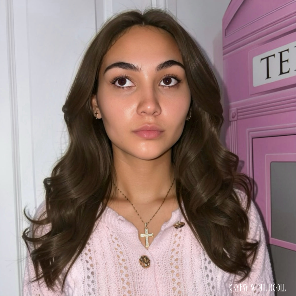 A woman with long, wavy brown hair is shown from the chest up, wearing a light pink knit cardigan with gold buttons and a gold cross pendant necklace. She is standing indoors in front of a white door and a pink telephone booth prop by Gypsy Soul Doll.