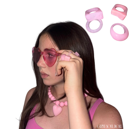 A woman with long dark hair models pink heart-shaped sunglasses, a pink beaded necklace with a heart pendant, and pink acrylic rings, shown against a plain white background with floating pink rings.