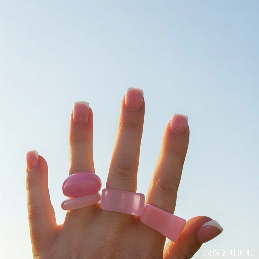 A hand with a French manicure wearing four chunky pink rings against a clear blue sky background.