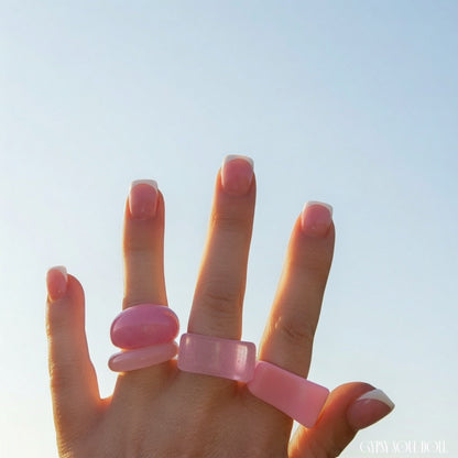 A hand with a French manicure wearing four chunky pink rings against a clear blue sky background.