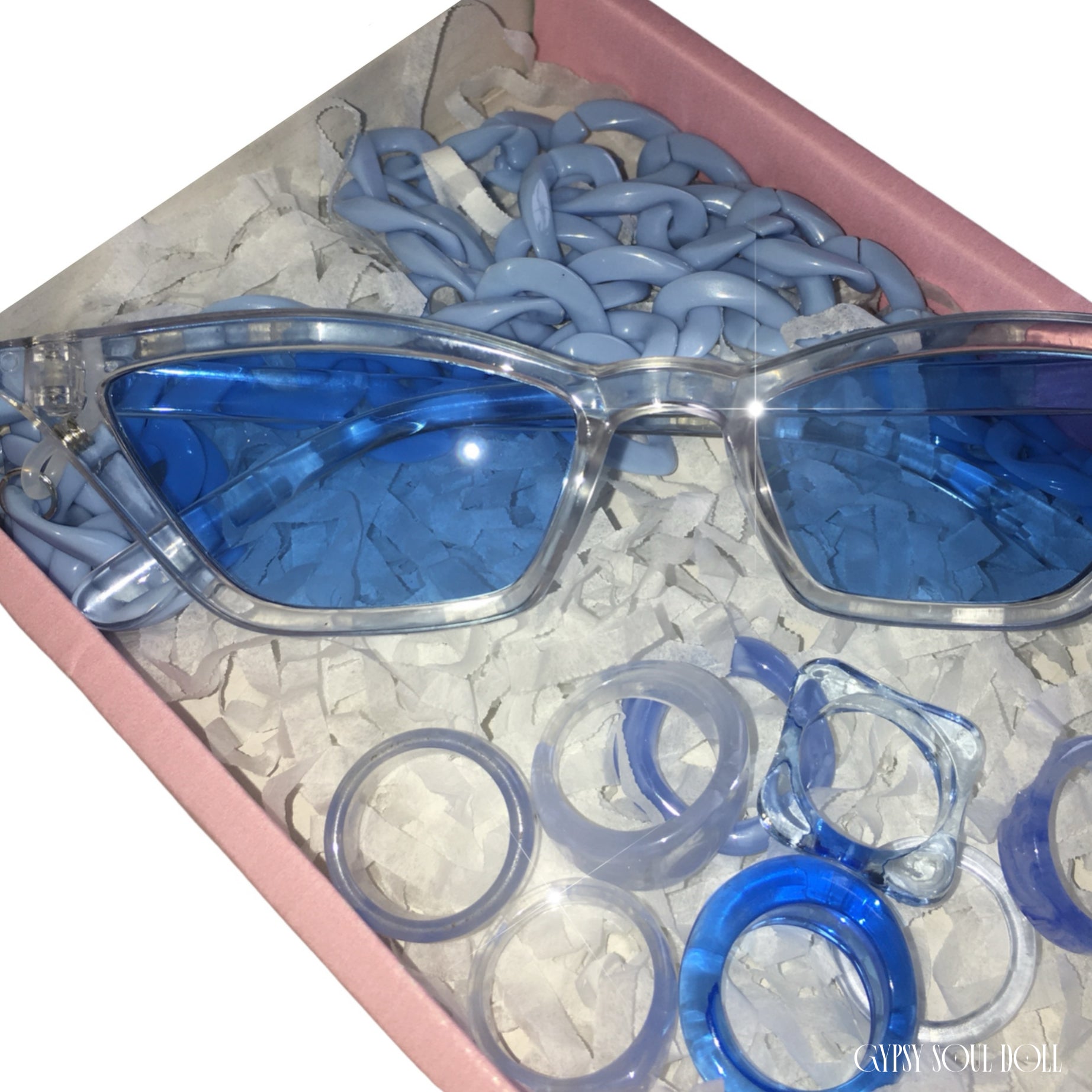 A pink Gypsy Soul Doll presentation box packed with white shredded paper, showcasing transparent cat-eye sunglasses with blue lenses, a powder blue acrylic link chain, and a curated stack of eight matching blue chunky resin rings.