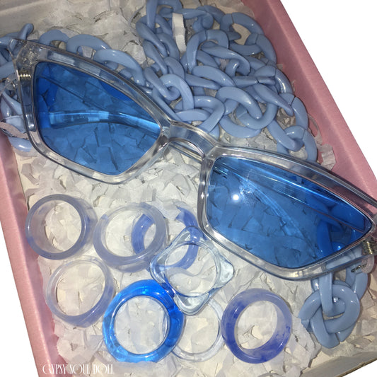A pink Gypsy Soul Doll presentation box packed with white shredded paper, showcasing transparent cat-eye fashion glasses with blue lenses, a powder blue acrylic link chain, and a curated stack of eight matching blue chunky resin rings.