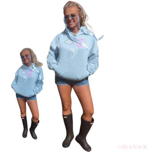 A dual-pose studio mockup of a blonde model showcasing the Gypsy Soul Doll baby blue graphic oversized street-style hoodie featuring pink and white evil eye, peace sign, and heart doodles, styled with denim shorts and black hunter festival wellington boots.