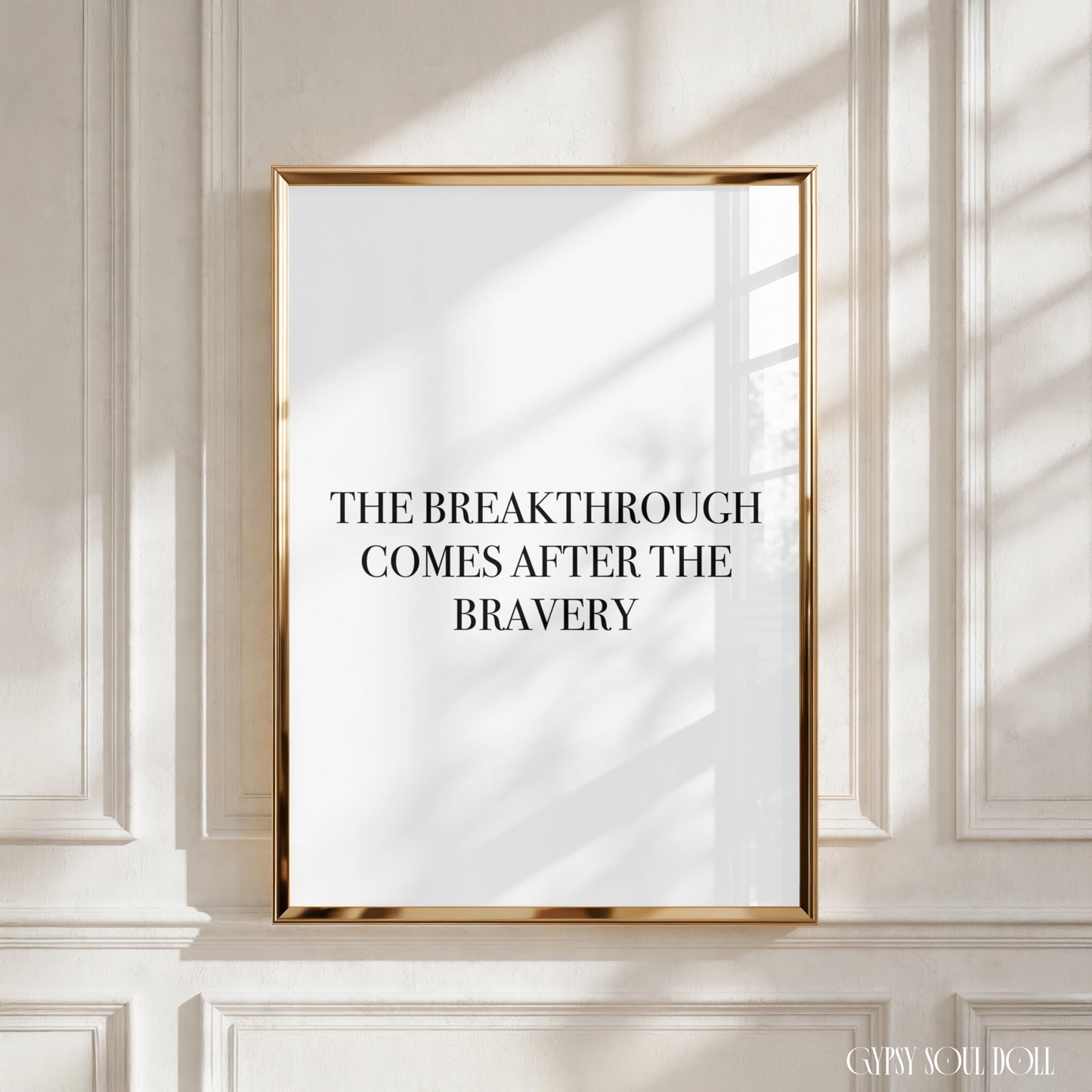 A framed typographic wall art print with a gold frame hangs on a white paneled wall. The print has a white background with black and burgundy text reading, ' The breakthrough comes after the bravery’.