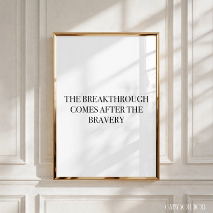 A framed typographic wall art print with a gold frame hangs on a white paneled wall. The print has a white background with black and burgundy text reading, ' The breakthrough comes after the bravery’.