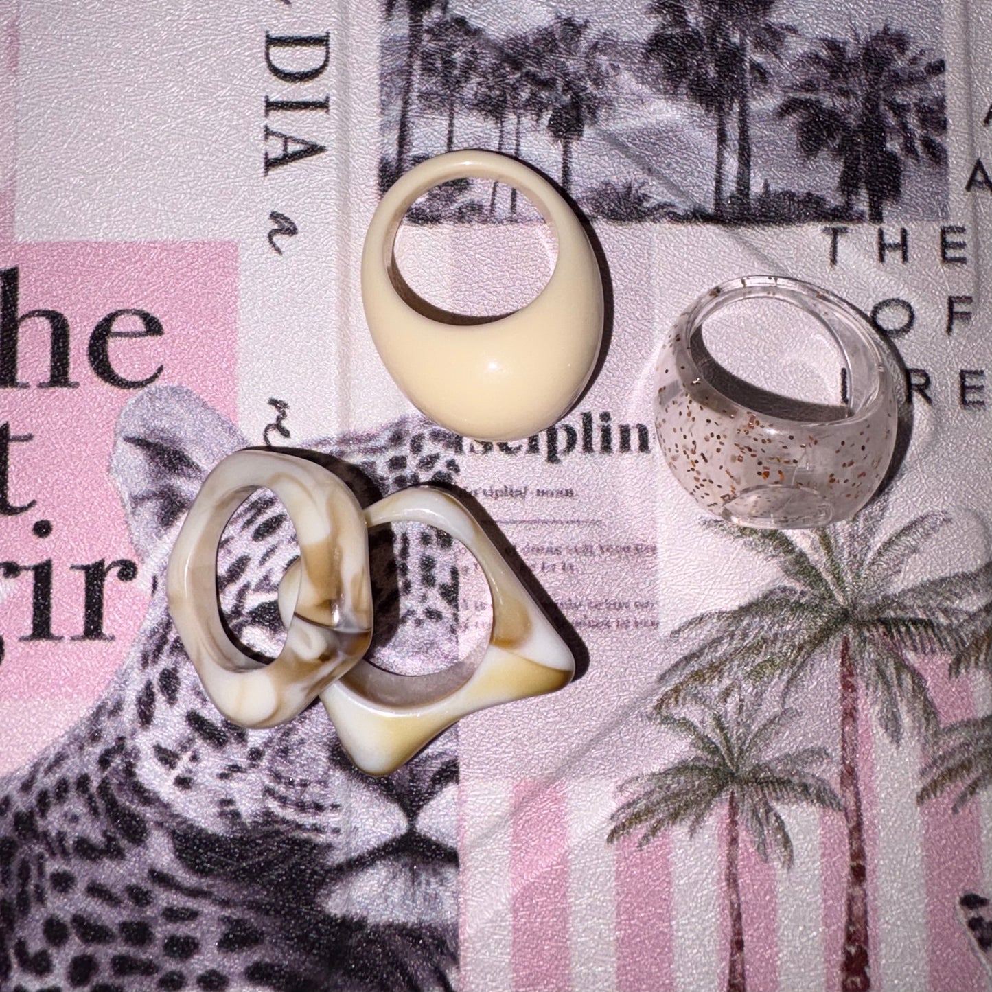Four sculptural rings arranged on a printed paper background with a leopard and palm tree print. The rings include one smooth cream oval ring, one pair of interlocking cream-and-brown abstract rings, and one clear ring with small gold speckles.