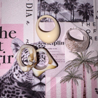 Four sculptural rings arranged on a printed paper background with a leopard and palm tree print. The rings include one smooth cream oval ring, one pair of interlocking cream-and-brown abstract rings, and one clear ring with small gold speckles.