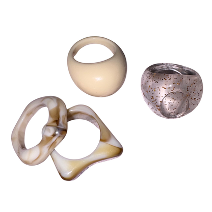 Four sculptural resin rings displayed on a solid white background. The set includes a chunky cream-colored ring with a loop, a smooth cream-colored bowl-shaped ring, and a translucent speckled brown ring.