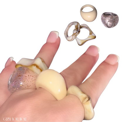 A hand with a classic French manicure modelling a four-piece stack of chunky Y2K-style acrylic rings in shades of cream, marbled brown beige, and translucent gold glitter, with a digital cutout of the rings floating above against a clean white background stamped with the Gypsy Soul Doll watermark.