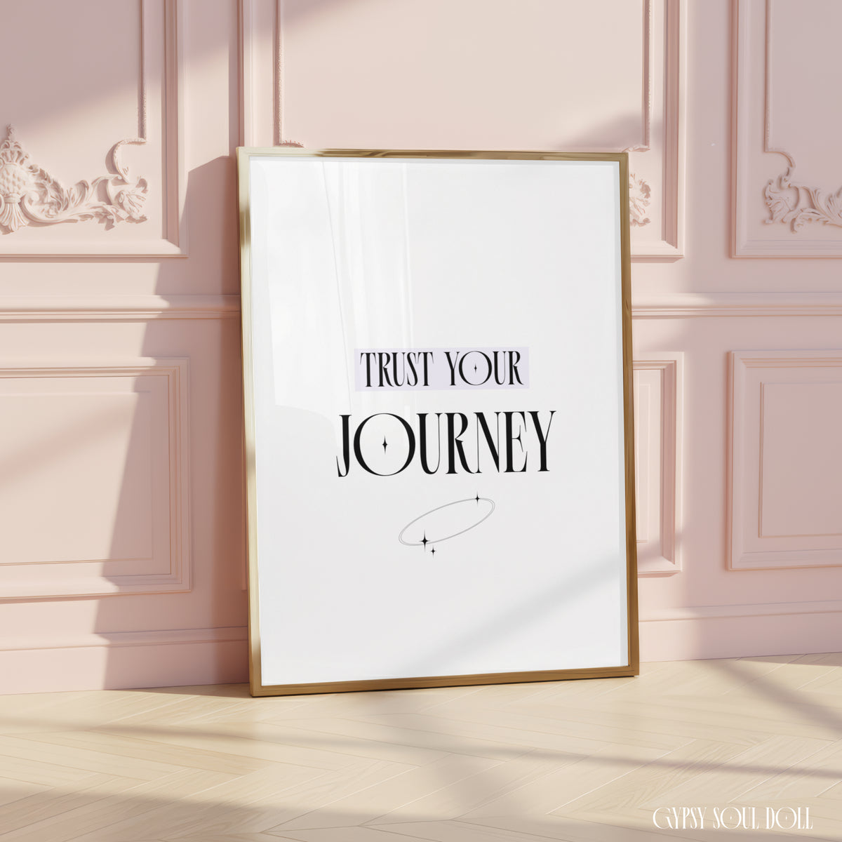 A framed typographic wall art print leaning against a pale pink panelled wall on a light wood floor. The print has a white background with the text 'TRUST YOUR JOURNEY' in black and a small minimalist orbit graphic by Gypsy Soul Doll.