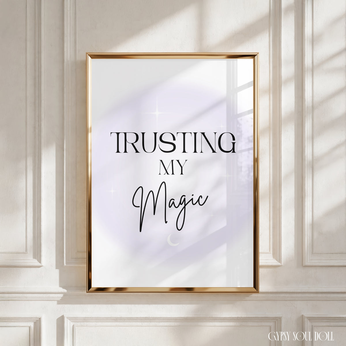 A framed art print with the text 'TRUSTING MY Magic' in black lettering on a white background with a soft lavender glow, set in a gold frame and leaning against a white panelled wall with soft window shadows by Gypsy Soul Doll.