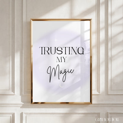 A framed art print with the text 'TRUSTING MY Magic' in black lettering on a white background with a soft lavender glow, set in a gold frame and leaning against a white panelled wall with soft window shadows by Gypsy Soul Doll.