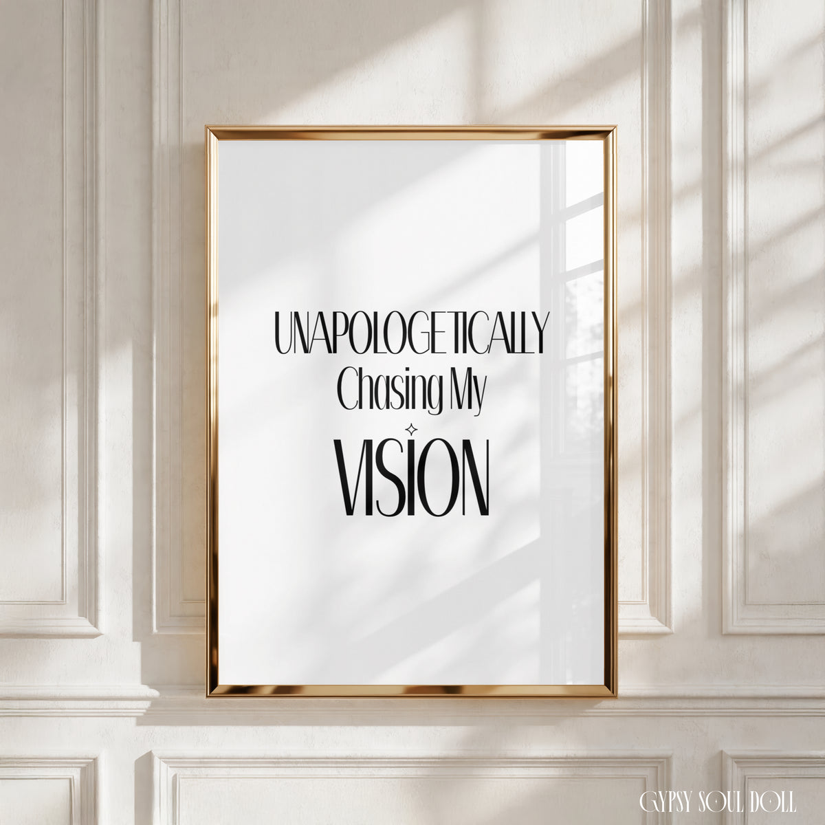 A framed typographic wall art print with the text 'UNAPOLOGETICALLY Chasing My VISION' in black serif lettering on a white background, set in a gold frame against an off-white paneled wall with soft window shadows by Gypsy Soul Doll.