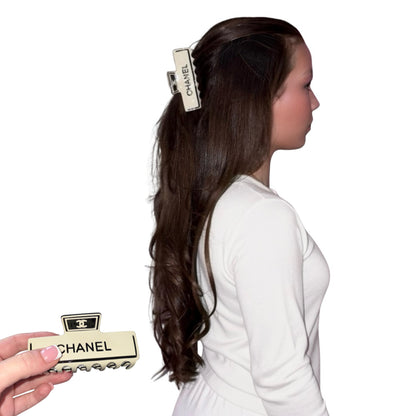 A model in a white loungewear outfit is shown in profile, wearing a large cream-coloured Chanel hair claw clip. A hand in the lower-left corner holds a second, clearer view of the same cream and black Chanel hair claw clip against a plain white background.