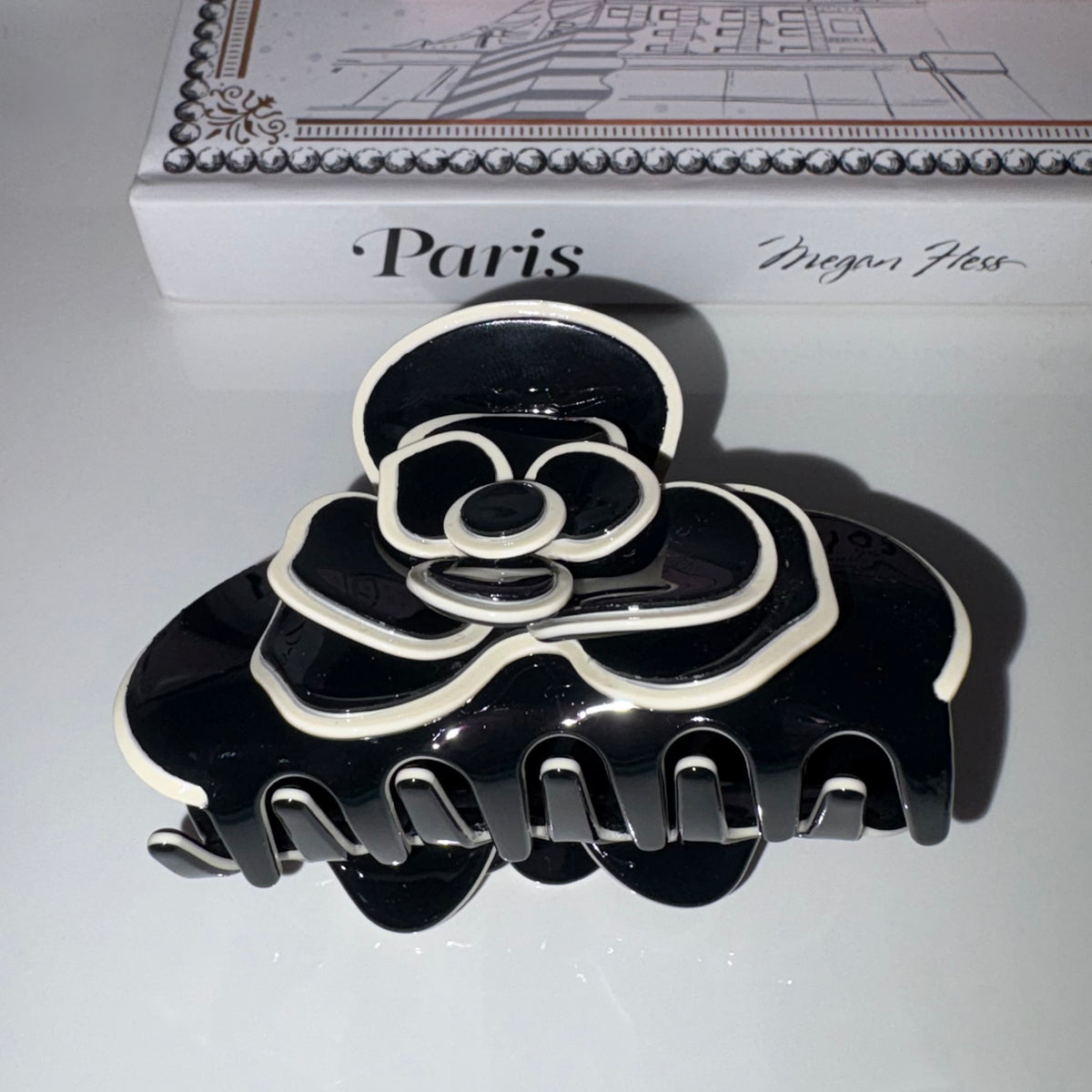 A glossy black hair claw clip with white outline detailing, styled in front of a white book with 'Paris' and 'Meghan Ness' printed on the cover.
