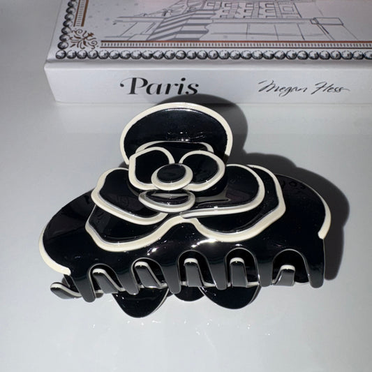 A glossy black hair claw clip with white outline detailing, styled in front of a white book with 'Paris' and 'Meghan Ness' printed on the cover.