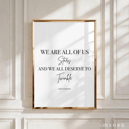 A framed typographic wall art print with a gold frame hangs on a white panelled wall. The print features black text reading, 'WE ARE ALL OF US Stars AND WE ALL DESERVE TO Twinkle' with 'Marilyn Monroe' printed below, and soft window shadows fall across the artwork and wall.
