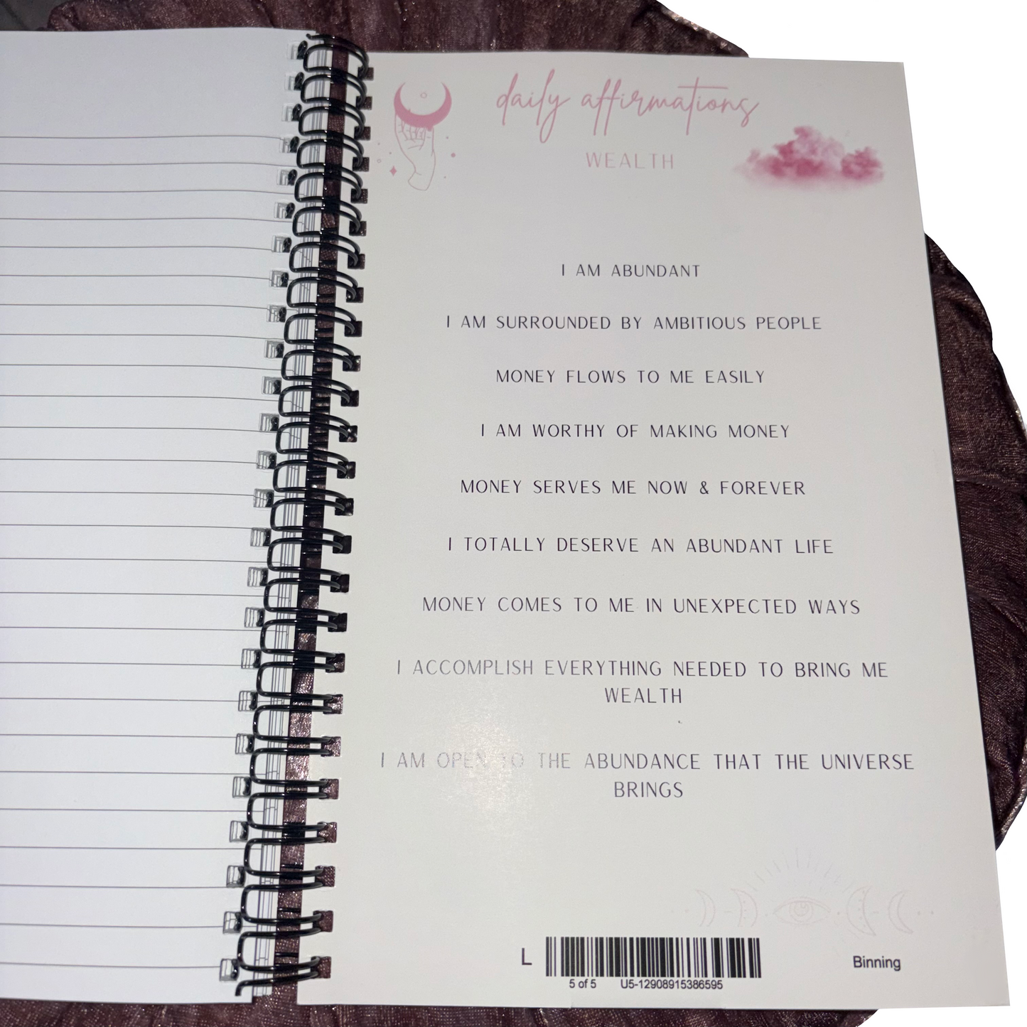 Open page of the spiral-bound notepad showcasing a list of positive wealth affirmations, pink cosmic moon graphics, and clean lined paper space.