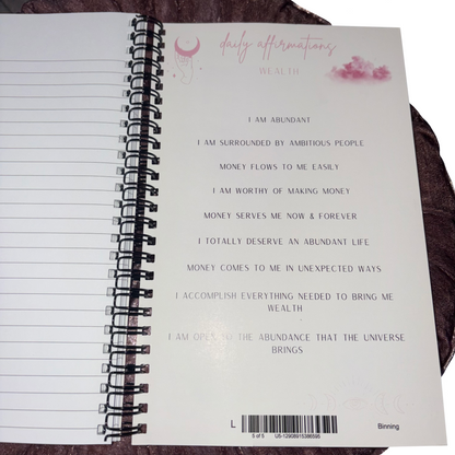 Open page of the spiral-bound notepad showcasing a list of positive wealth affirmations, pink cosmic moon graphics, and clean lined paper space.