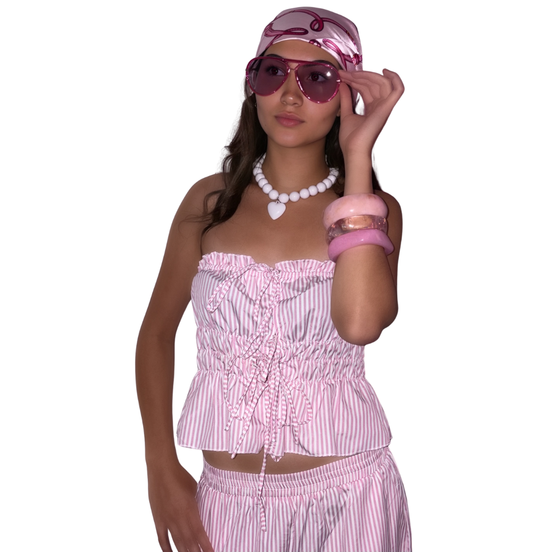 A woman modelling a pink and white striped two-piece set consisting of a strapless ruffled crop top and matching trousers, accessorised with a matching pink and white boho head wrap, pink sunglasses, a white chunky retro beaded necklace, and pink bracelets against a plain white background.