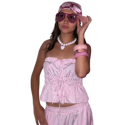 A woman modelling a pink and white striped two-piece set consisting of a strapless ruffled crop top and matching trousers, accessorised with a matching pink and white boho head wrap, pink sunglasses, a white chunky retro beaded necklace, and pink bracelets against a plain white background.