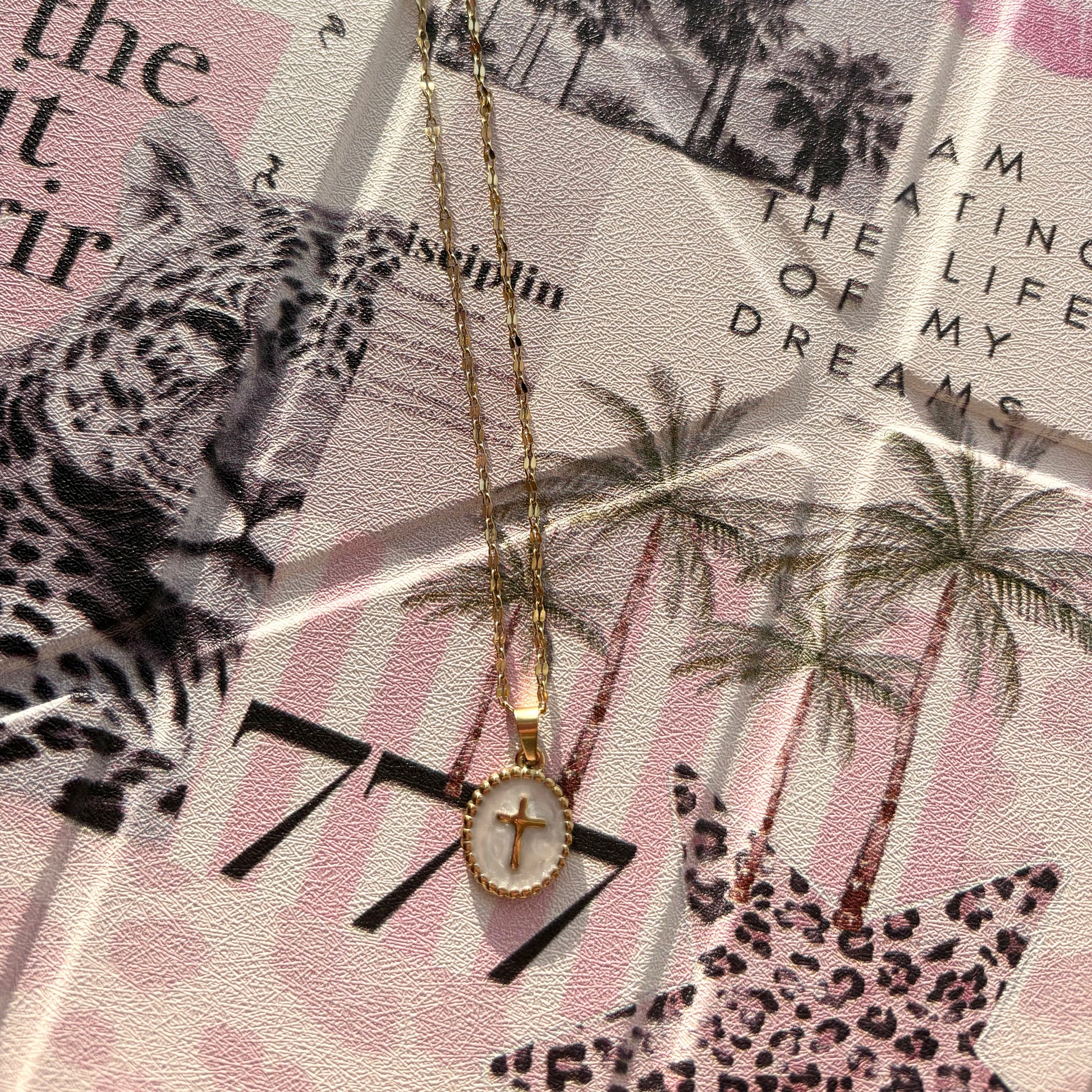 A gold-plated cross pendant necklace with a white oval inlay, displayed on a styled collage background featuring a leopard print, palm trees, and text.