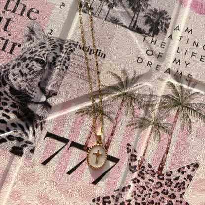 A gold-plated cross pendant necklace with a white oval inlay, displayed on a styled collage background featuring a leopard print, palm trees, and text.