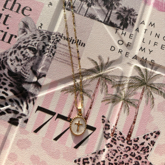 A gold-plated cross pendant necklace with a white oval inlay, displayed on a styled collage background featuring a leopard print, palm trees, and text.