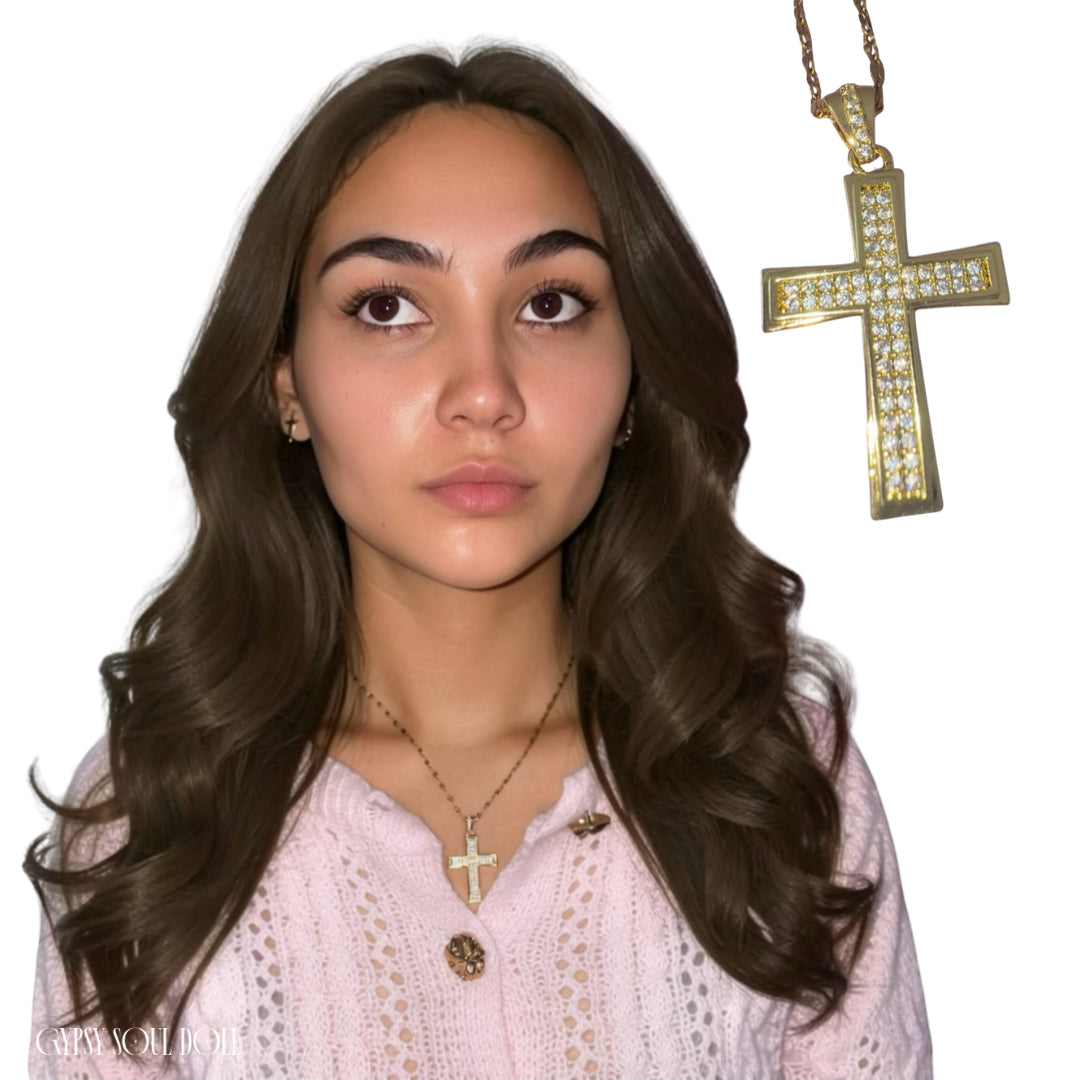 A model with long wavy brunette hair wearing a pink lace cardigan and a gold iced cross pendant necklace.