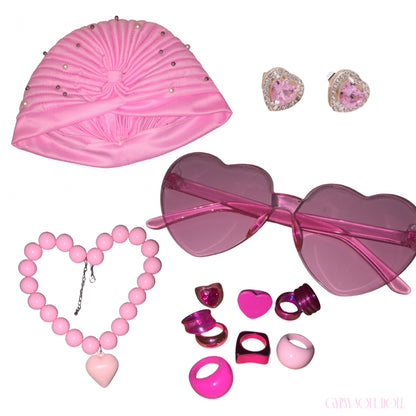 A pink themed accessory set on a white background, including a pink satin headband with rhinestones, heart-shaped fashion glasses, a pink beaded heart necklace, heart stud earrings, and various pink and chunky rings.