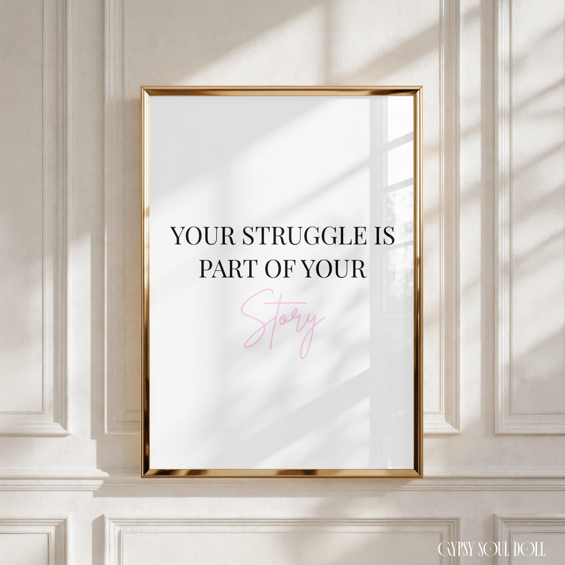 A framed motivational art print with the text 'YOUR STRUGGLE IS PART OF YOUR Story' in black and pink lettering on a white background, enclosed in a thin gold frame and mounted on a white paneled wall with soft window shadow patterns.