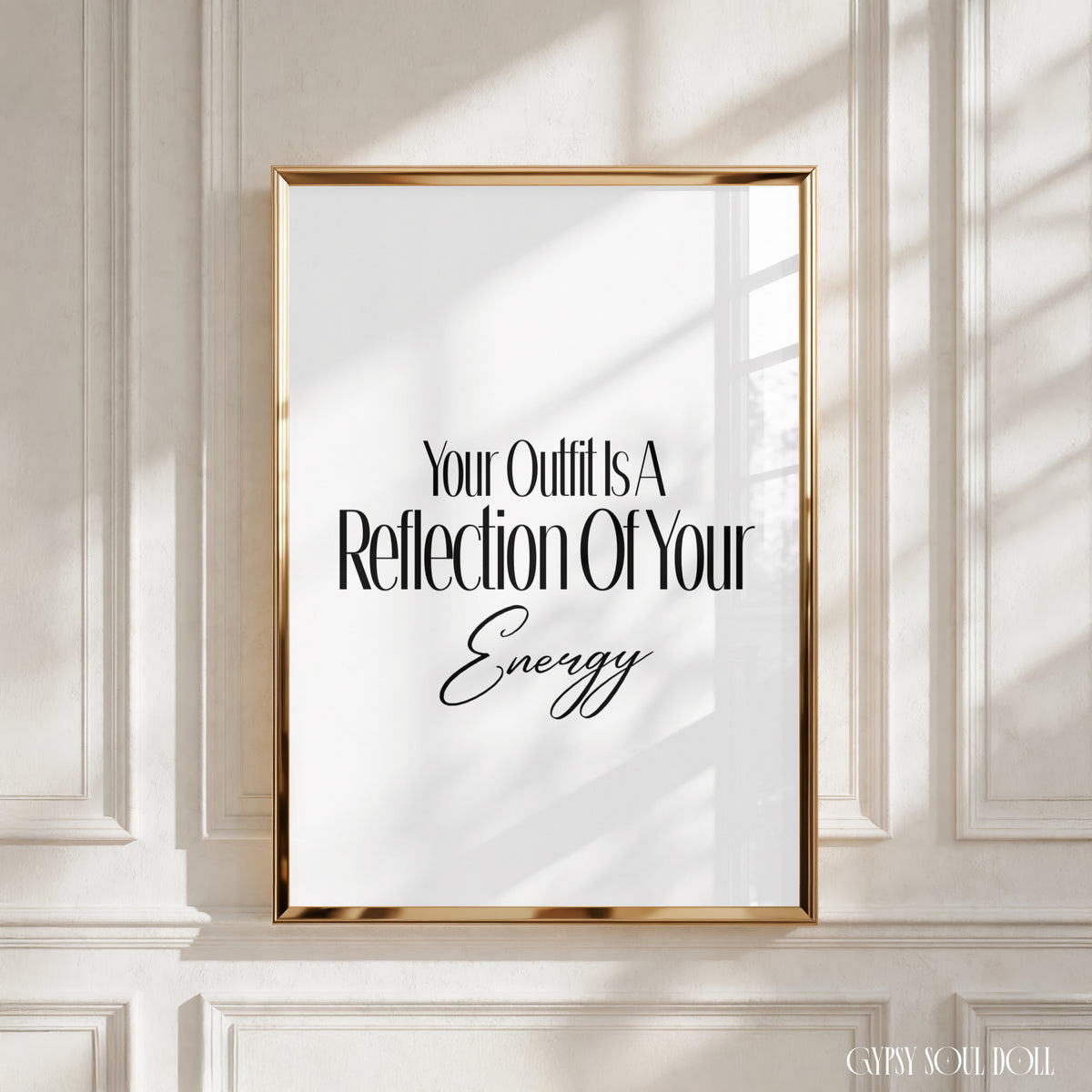 A framed typographic wall art print with the phrase 'Your Outfit Is A Reflection Of Your Energy' in black text on a white background, set in a gold frame and displayed against an off-white panelled wall with soft window shadows by Gypsy Soul Doll.